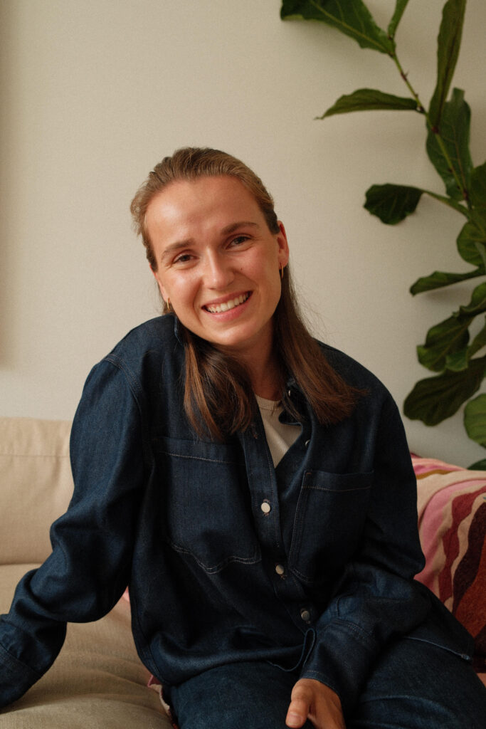 Image of Flip sitting on a couch with a plant behind her. She is smiling and wearing a denim jacket.